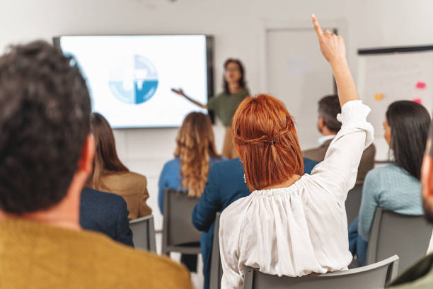 Red-haired Woman Raising Hand during Business Presentation Businesswoman with red hair raising hand to ask question during a SWOT analysis presentation in large office room. Diverse audience, viewed from back. teacher projector stock pictures, royalty-free photos & images