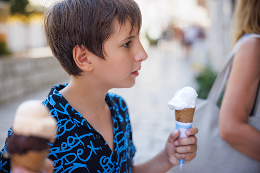 Preadolescent boy holding ice cream served on scoops in biscuit cone, taking a walk with his family in the street