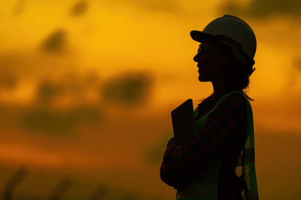 Half body silhouette of female engineers smiling and holding laptop stand near the industrial plant The twilight sky resembles a sunset, glowing with golden light, wearing a helmet and vest. Half body silhouette of male and female engineers smiling and holding laptop stand near the industrial plant The twilight sky resembles a sunset, glowing with golden light, wearing a helmet and vest. factory worker silhouette stock pictures, royalty-free photos & images