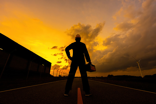 Silhouette of the back of a male engineer stand near the industrial plant holding a toolbox wearing a mechanic's suit The twilight sky resembles a sunset, golden light, wears a helmet.
