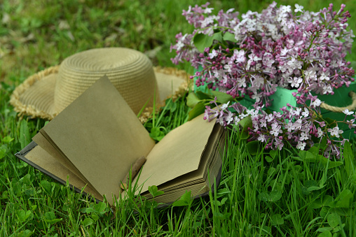 Vintage still life with open book or diary, hat and lilac flowers bouquet in the garden. Summer and spring season concept