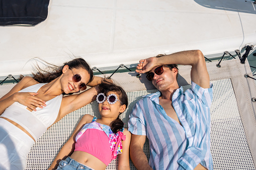 Caucasian attractive family lying down on deck of yacht while yachting. Young beautiful couple hanging out and spend time with daughter while catamaran boat sailing during holiday summer trip together