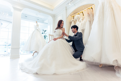 Asian beautiful bride trying on wedding gown with her fiance in studio. Attractive young couple feeling happy and relax, enjoying preparation for marriage ceremony in fitting room at the bridal store.