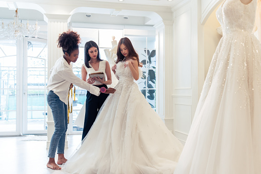 Beautiful bride discussing details with the wedding planner in studio. Attractive bridal shop owner women use digital tablet help customer choosing wedding gown at the store. Marriage ceremony concept