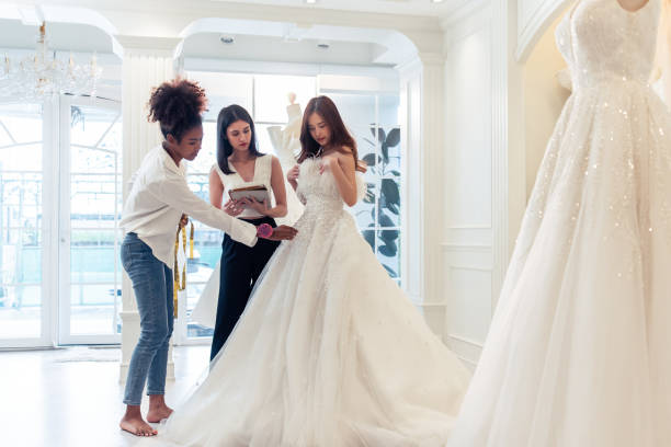 beautiful bride discussing details with the wedding planner in studio. attractive bridal shop owner women use digital tablet help customer choosing wedding gown at the store. marriage ceremony concept - bride wedding fashion evening gown imagens e fotografias de stock