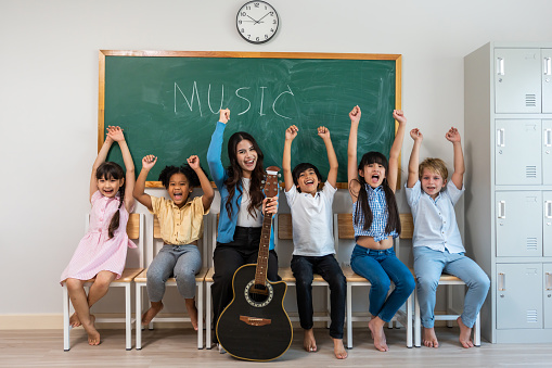 Portrait of diverse children student in classroom at elementary school. Attractive female instructor teacher sitting with young kid after teaching a music subject lesson with happiness at kindergarten