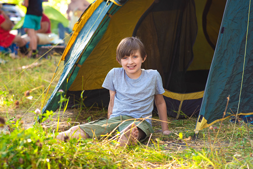a little boy sits on the grass in front of a tent at a camp in the forest