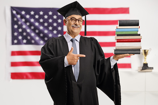 Mature man in a graduation gown holding a pile of books and pointing in front of the USA flag