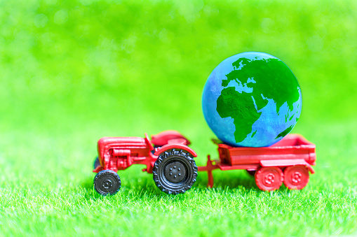Toy tractor carries a globe across a lush green meadow. Creative concept related to sustainable agriculture, environmentally-friendly and responsible methods in farming and cultivating crops.