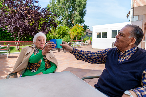 Two elderly individuals, a man and a woman, sit in a beautifully landscaped garden at an elderly care home, happily raising vibrant glasses to celebrate, bathed in sunlight.