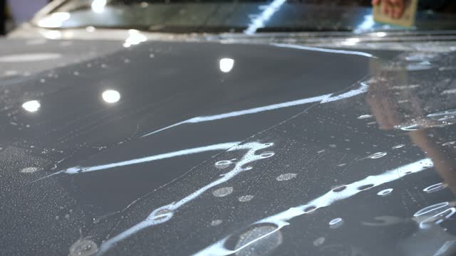 A hand with a plastic scraper glues a protective film on the new car hood.