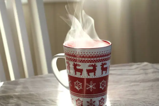 Photo of Steaming Cup of Coffee or Tea on Table. White linen Tablecloth. Close up ceramic red Christmas mug with smoke on old table in dining room interior background. Hot Drink Concept. Cozy Morning.