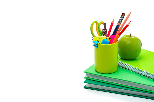 Multicolored school or office supplies in a desk organizer and green books isolated on white background. The composition is at the right of an horizontal frame leaving useful copy space for text and/or logo at the left. High resolution 42Mp studio digital capture taken with Sony A7rII and Sony FE 90mm f2.8 macro G OSS lens