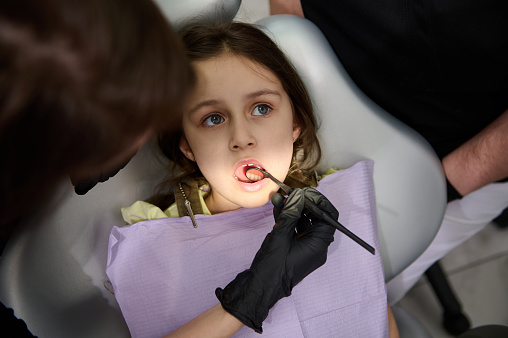 Close-up Caucasian little child girl with open mouth, is being examined by a dental hygienist, dentist doctor in pediatric dentistry clinic. Dental light directed at her oral cavity. Dental practice