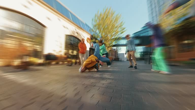 Trendy Zoom Blur Edit of Young B-boy Breakdancing On City Street Among Modern Buildings In Urban Area. Group Of Friends Cheering On Background, Supporting Breakdancer Practising Choreography.