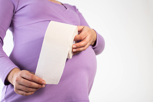 A pregnant girl in a lilac blouse holds toilet paper against the background of her belly. The concept of constipation in pregnant women.