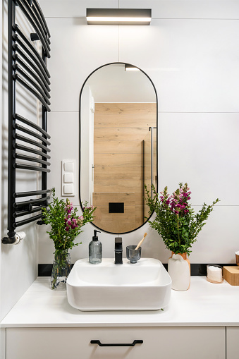 modern interior in home bathroom, washbasin with black faucet, under sink drawer, ceramic vase with flowers and mirror with picture on tiled wall