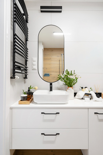 light interior in elegant bathroom, ceramic washbasin with black mixer, vase with blossom flowers, cosmetics products in metal basket and mirror on tiled wall