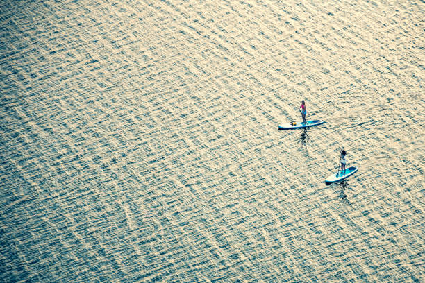 Trakai, Lithuania - June 21 2023: Two paddleboarders girls boarding or floating on a stand up paddle on the quite water of a lake with warm summer sunset colours, water sport, aerial view