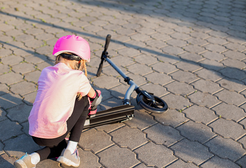 A 7-year-old girl in a pink helmet and protective gear fell off a scooter. The concept of safe riding a scooter and a bicycle without injury.