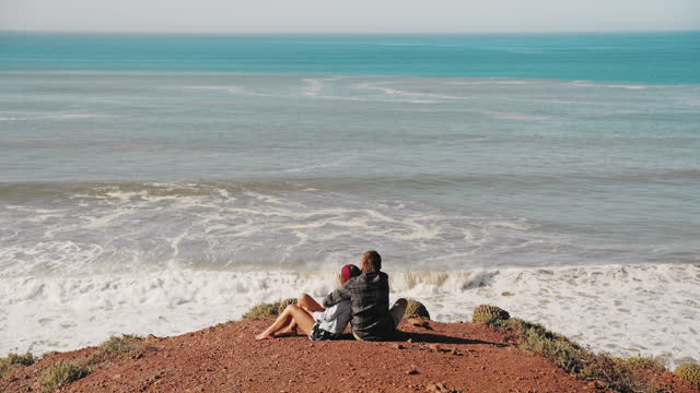 Couple embracing and looking at seascape while sitting on cliff at Morocco