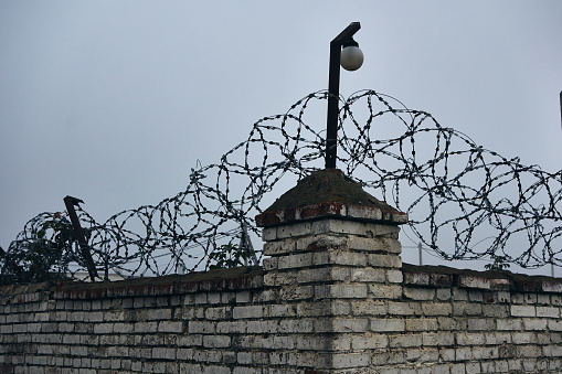 A fence made of bricks, a spiral barbed wire is stretched from above, a lighting lamp hangs on a metal bracket, cloudy and foggy, depressing mood