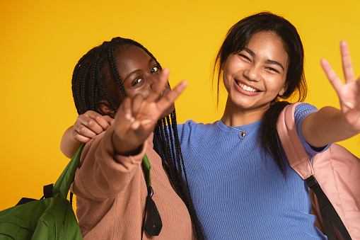 Two teenage girls embracing each other and making peace sign against yellow background