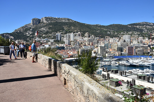 Monaco-Ville, Monaco - April 24, 2023: Tourists Standing, Sitting Down, Talking To One Another, Taking Picture Of Monaco Harbor, Mountain, Docked Recreational Boat And Skyline Of Monaco During Springtime