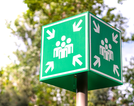 A bright green emergency evacuation sign with a directional arrow and a running figure hanging from the ceiling in the hallway. Selective focus. Close-up