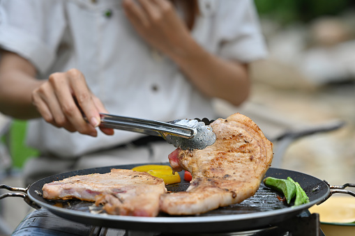 Close up and cropped image with female camper grilling pork chop on pan with picnic gas stove, Fine dining outdoor concept.
