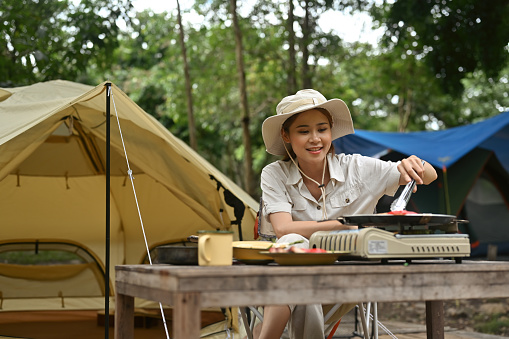 Portrait with Young Asian female camper in safari clothes cooking meal in front of camping tent, Fine dining and outdoor concept.