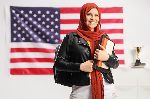 Female student with a hijab holding books and looking at camera in front of a USA flag