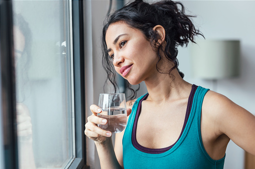 Shot of beautiful sporty woman drinking a glass of water while standing near the window at home