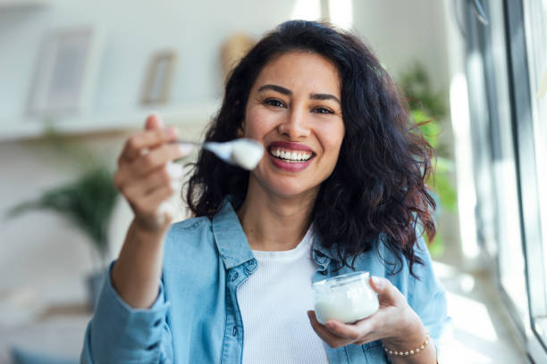 istock Beautiful smiling woman having fun while feeding yogurt to camera at home 1504173548