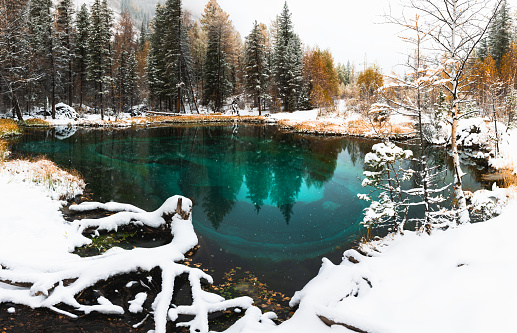 Blue geyser lake in autumn forest during snowfall. Altai mountains, Siberia, Russia. Lake with blue water and white snow. Autumn landscape