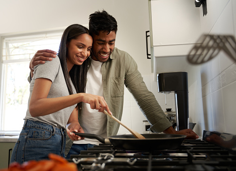 Happy young biracial couple wearing casual clothing cooking food in saucepan over stove in kitchen