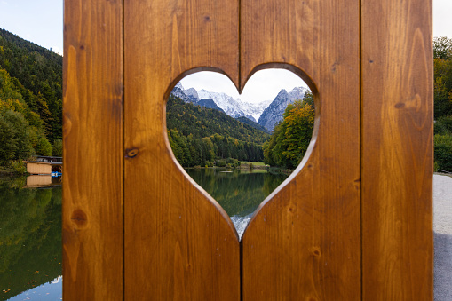 A medium shot of a mountainous landscape reflecting over a lake in Garmisch, Germany. It is shot through a heart carved in a wooden board.