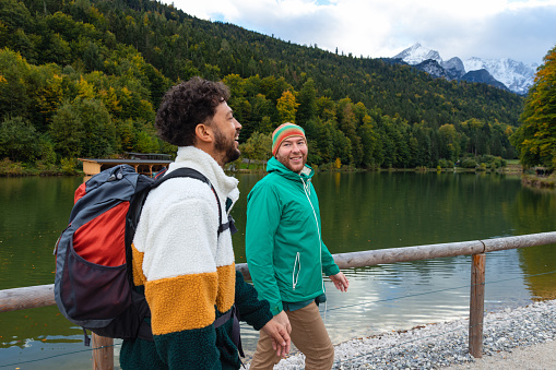 A medium shot of a  multi-ethnic homosexual couple walking together besides a lake with a snow capped mountainous landscape in the background in Garmisch, Germany.