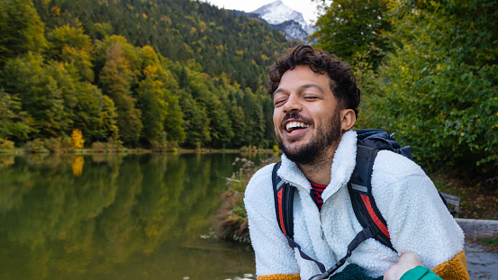A medium shot of a  multi-racial, mid-adult man laughing while he stands besides a lake with a snow capped mountainous landscape in the background in Garmisch, Germany.