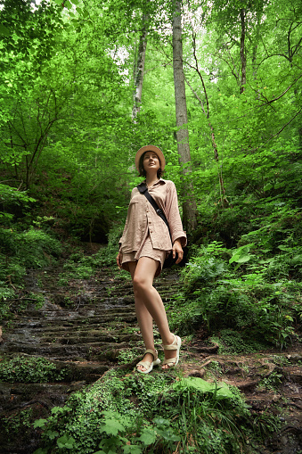 woman in a hat standing on a path in the forest, in the style of dark beige and green