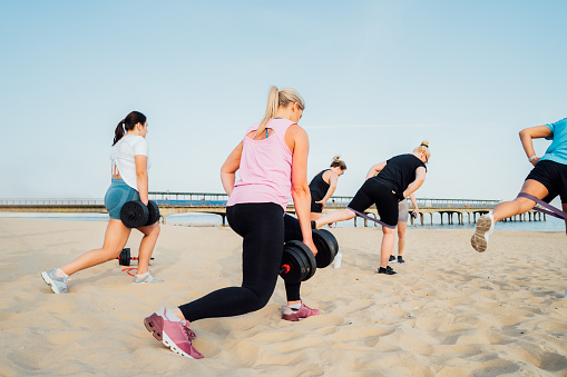 Back view women of various ages doing fitness workouts in class exercise with coach on the beach. Ladies working with dumbbells and resistance bands. Sport for health and wellbeing. Active lifestyle