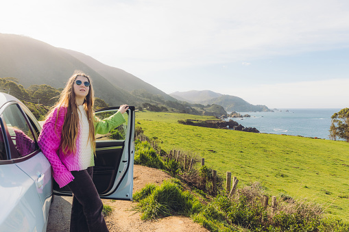 Smiling  female with long hair in colourful sweater contemplating road trip by car through the scenic coast if Big Sur, looking at the beautiful ocean and green hills in Monterey peninsula, United States