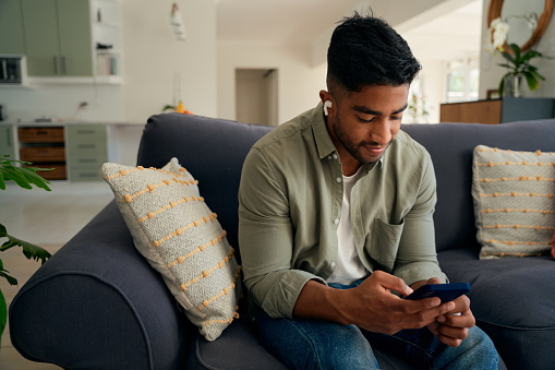 Young biracial man wearing casual clothing sitting on sofa with mobile phone and wireless earphones at home