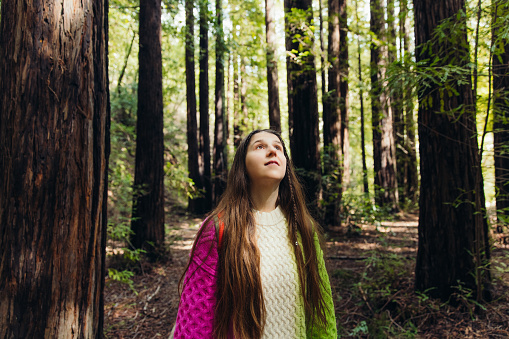 Portrait of female with long hair and backpack wearing colorful sweater walking in scenic forest with redwood trees in Monterey Peninsula. the USA
