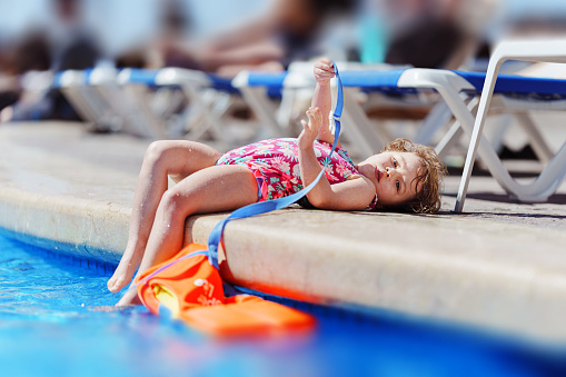 3 year old, multiracial girl sunbathes by the pool with her toes dipped in the water, while she looks tiredly at the camera.