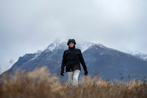 A senior woman walks on a cold day towards the camera in the middle of a field with spikes with a snowy mountain behind.