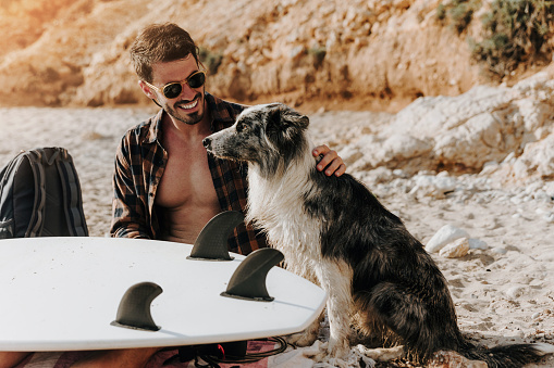 Young male surfer and his loyal dog enjoying the sun on a sandy beach and preparing their surfboard for a big waves. Perfect sunny day.