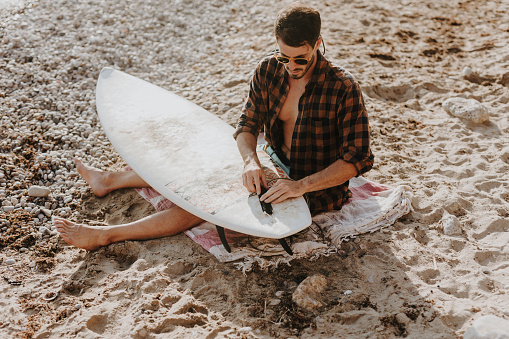Handsome male surfer enjoying on sunny beach while preparing surfboard for a next exciting wave ride. Summer leisure activity concept.