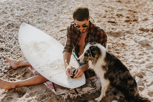 Young male surfer and his loyal dog enjoying the sun on a sandy beach and preparing their surfboard for a big waves. Perfect sunny day.