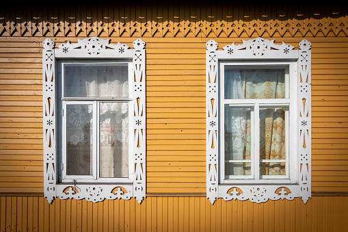 Two windows framed with carved wooden jambs decorated with traditional white patterns on the facade of a yellow painted Russian isba, a wooden-built house in northern Russia